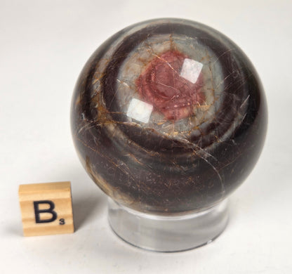 Polished Cherry Creek jasper sphere in various shades of red, orange, yellow, and green, with acrylic holder and a scrabble tile on a white background