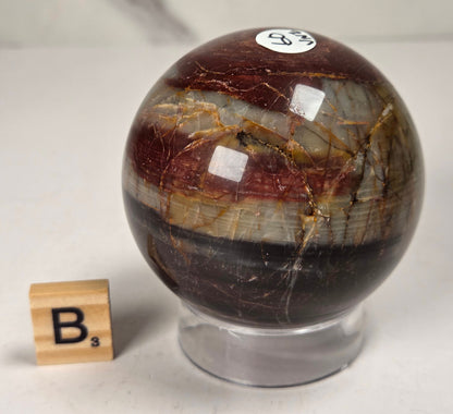 Polished Cherry Creek jasper sphere in various shades of red, orange, yellow, and green, with acrylic holder and a scrabble tile on a white background