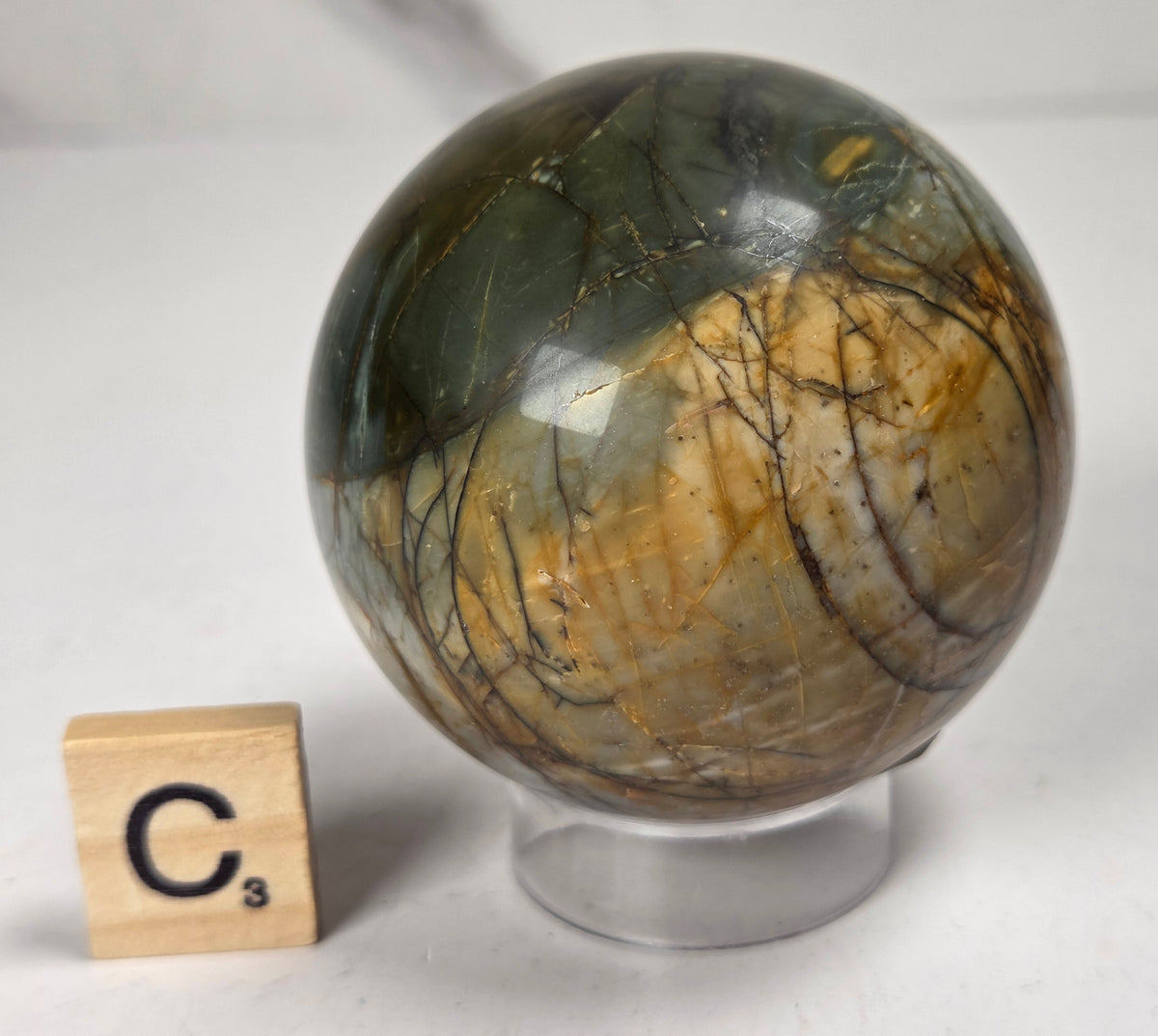 Polished Cherry Creek jasper sphere in various shades of red, orange, yellow, and green, with acrylic holder and a scrabble tile on a white background