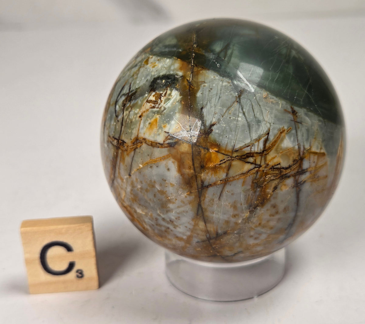 Polished Cherry Creek jasper sphere in various shades of red, orange, yellow, and green, with acrylic holder and a scrabble tile on a white background