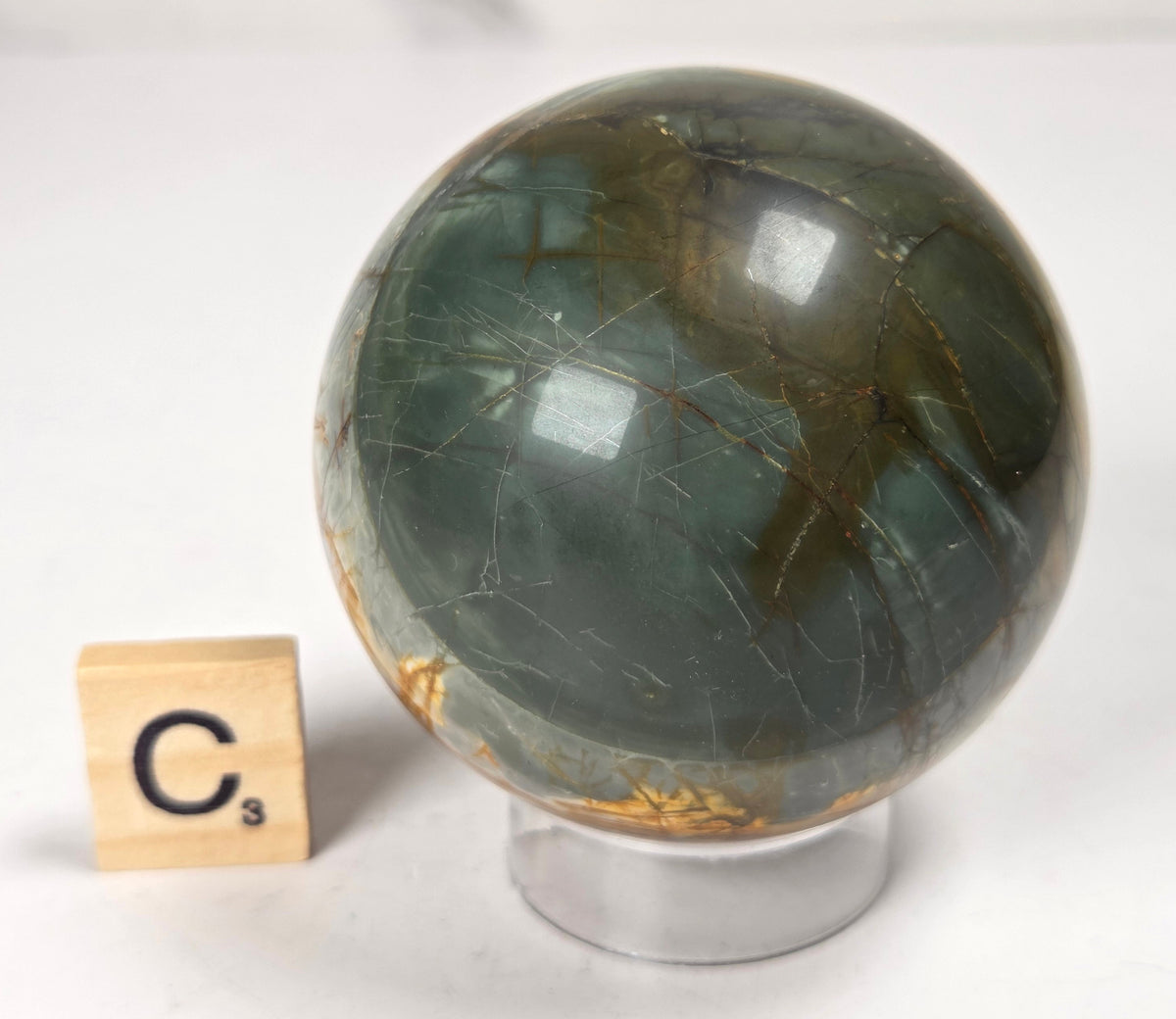 Polished Cherry Creek jasper sphere in various shades of red, orange, yellow, and green, with acrylic holder and a scrabble tile on a white background