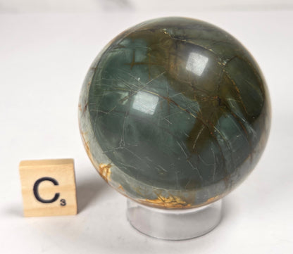 Polished Cherry Creek jasper sphere in various shades of red, orange, yellow, and green, with acrylic holder and a scrabble tile on a white background