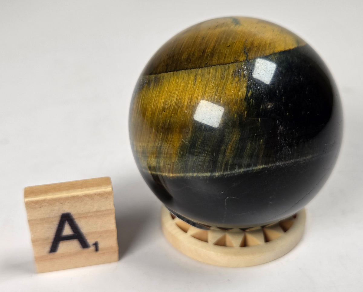 Blue Tigers Eye polished sphere carving with blue and yellow coloring on a wood stand with a scrabble tile against a white background