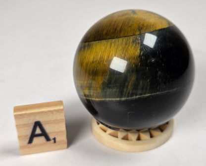 Blue Tigers Eye polished sphere carving with blue and yellow coloring on a wood stand with a scrabble tile against a white background