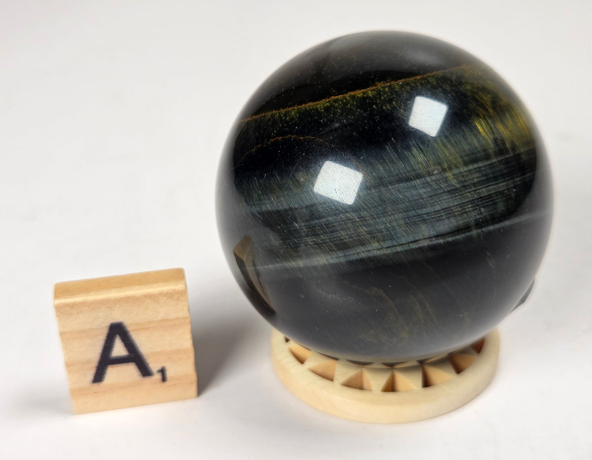 Blue Tigers Eye polished sphere carving with blue and yellow coloring on a wood stand with a scrabble tile against a white background