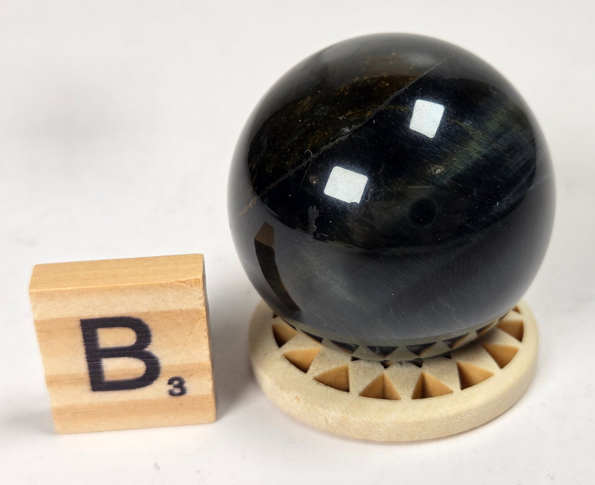 Blue Tigers Eye polished sphere carving with blue and yellow coloring on a wood stand with a scrabble tile against a white background