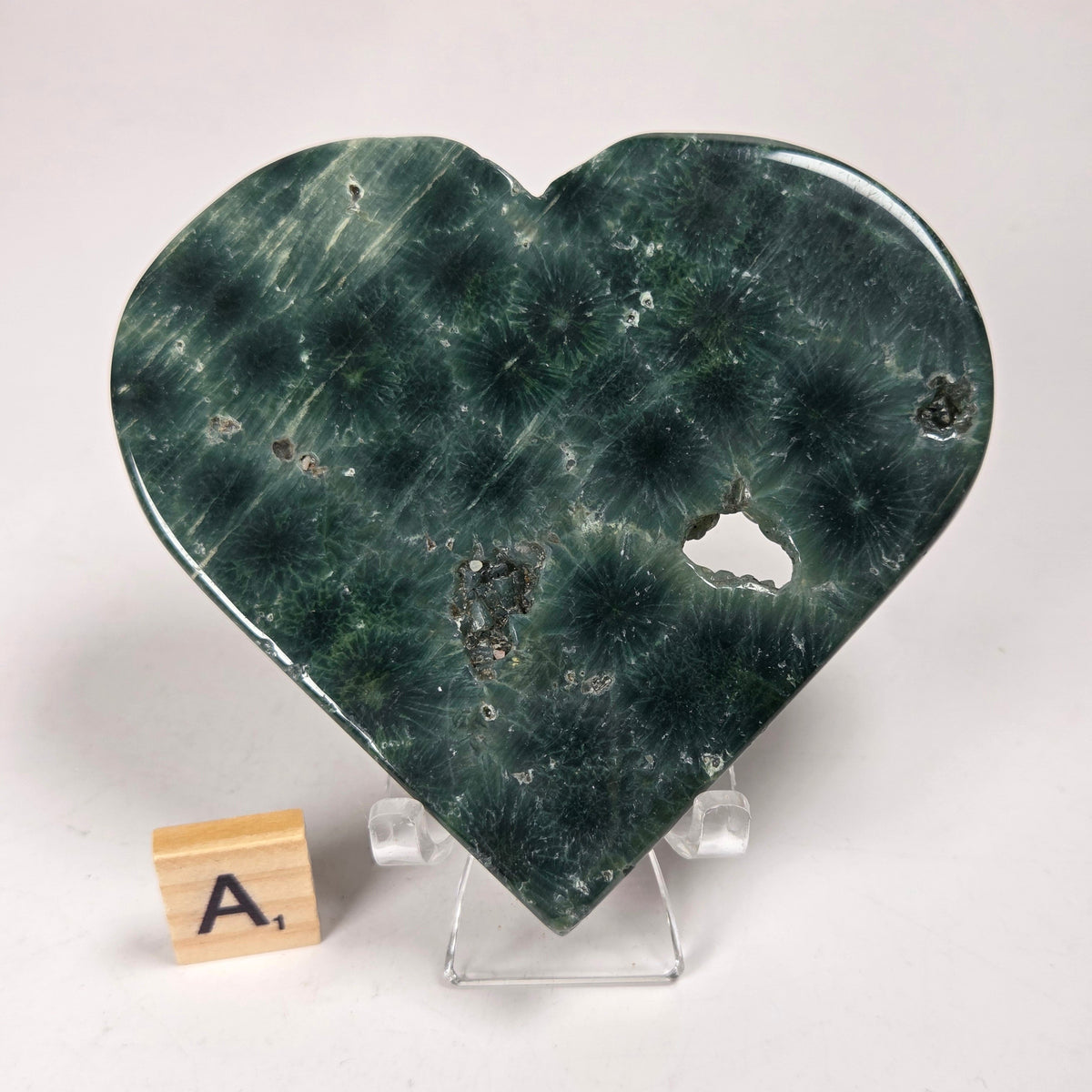 Green polished crystal heart carving with patterns and open spots on an acrylic chair with a scrabble tile against a white background
