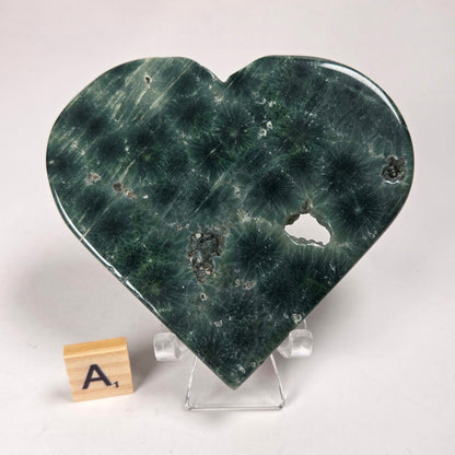 Green polished crystal heart carving with patterns and open spots on an acrylic chair with a scrabble tile against a white background