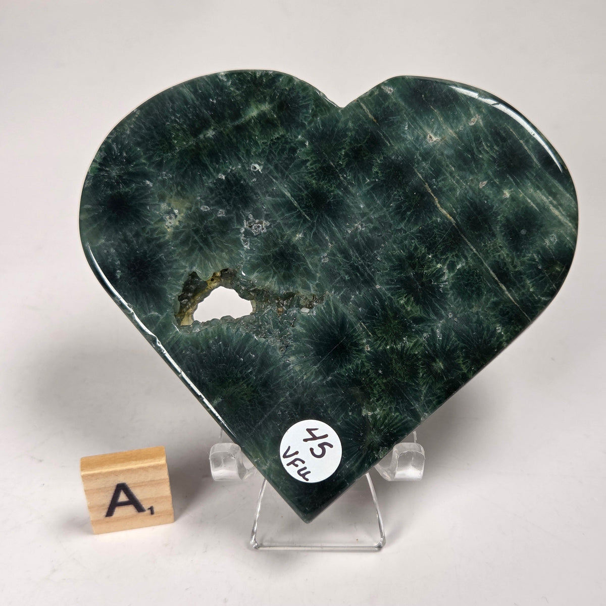 Green polished crystal heart carving with patterns and open spots on an acrylic chair with a scrabble tile against a white background