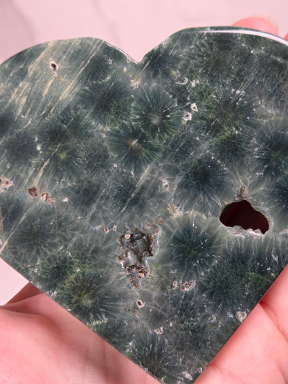 Close-up of a Green polished crystal heart carving with patterns and open spots being held in a hand against a white background