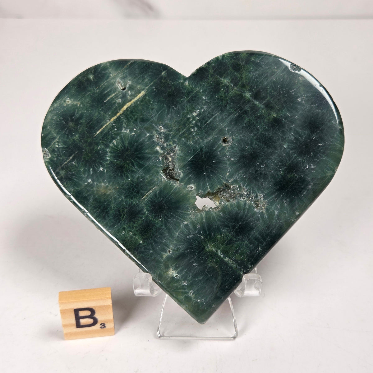 Green polished crystal heart carving with patterns and open spots on an acrylic chair with a scrabble tile against a white background