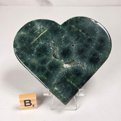 Green polished crystal heart carving with patterns and open spots on an acrylic chair with a scrabble tile against a white background