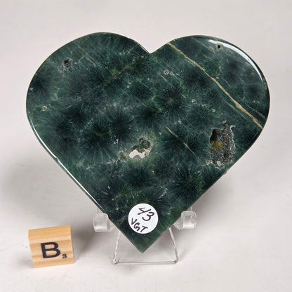 Green polished crystal heart carving with patterns and open spots on an acrylic chair with a scrabble tile against a white background