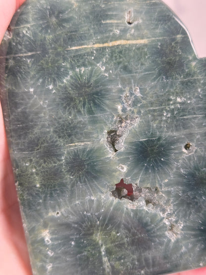 Close-up of a green polished crystal heart carving with patterns and open spots being held in a hand against a white background