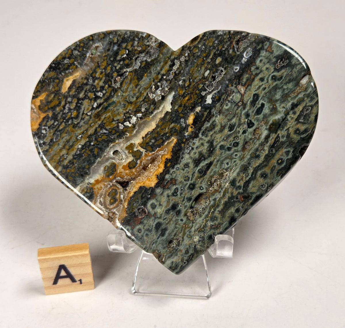 Polished crystal heart carving with green, yellow and white patterns on an acrylic chair with a scrabble tile against a white background