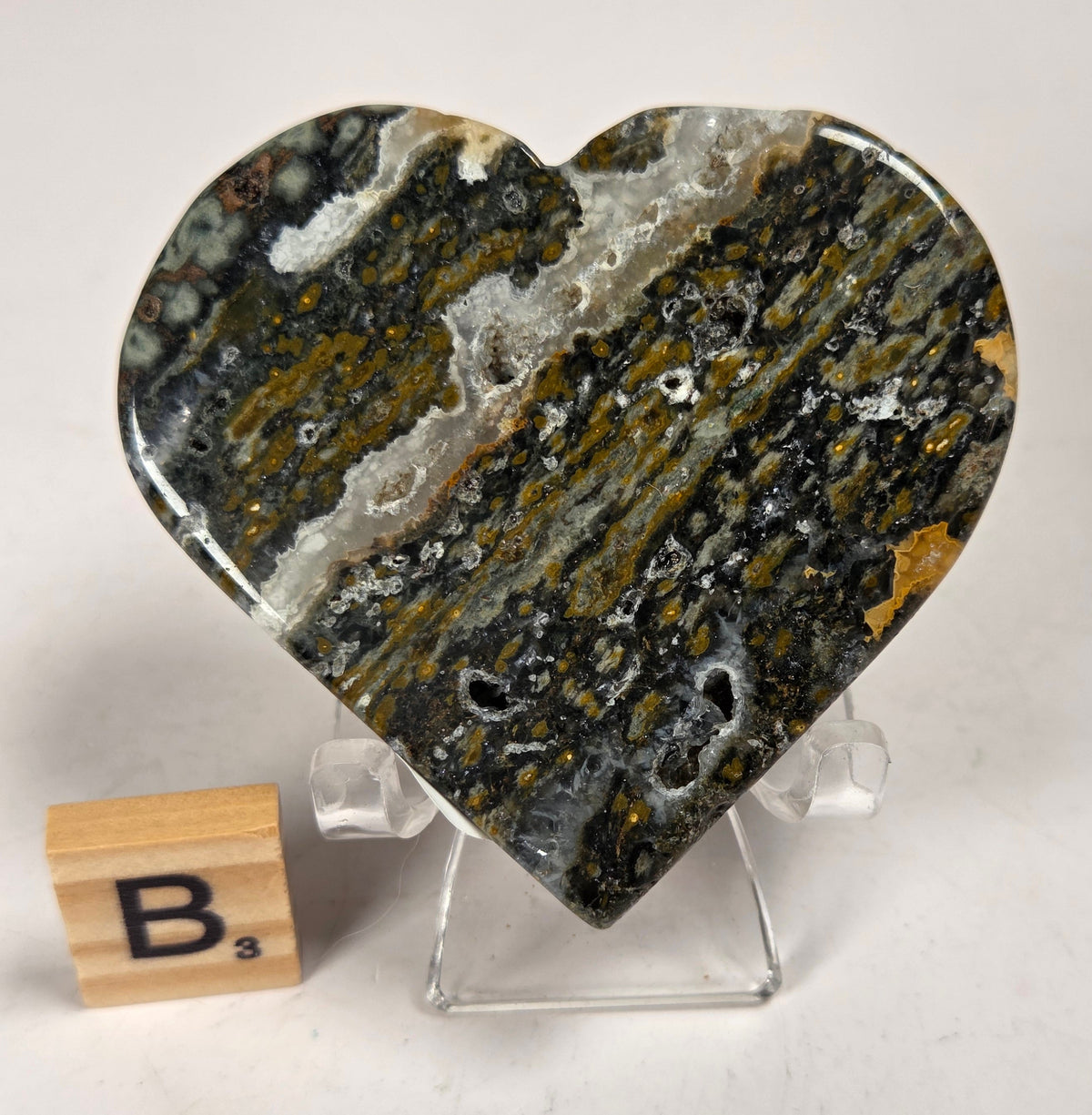 Polished crystal heart carving with green, yellow and white patterns on an acrylic chair with a scrabble tile against a white background