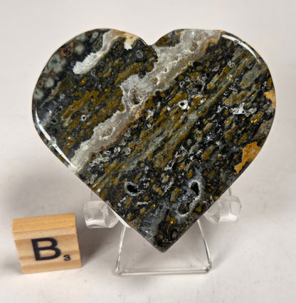 Polished crystal heart carving with green, yellow and white patterns on an acrylic chair with a scrabble tile against a white background