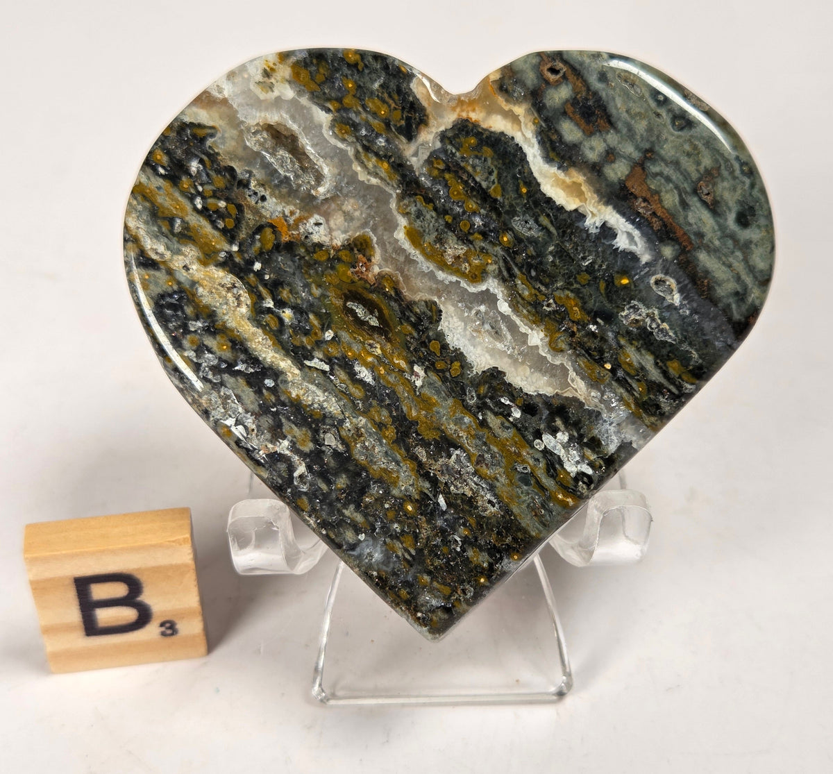 Polished crystal heart carving with green, yellow and white patterns on an acrylic chair with a scrabble tile against a white background