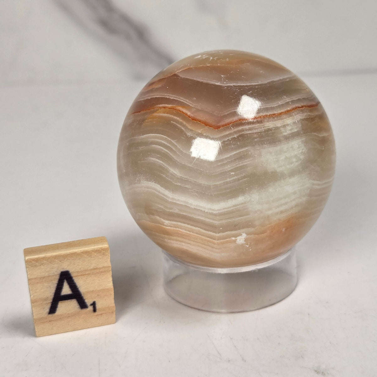 banded calcite spheres with green, brown and red banding on an acrylic stand with a scrabble tile against a marbled background