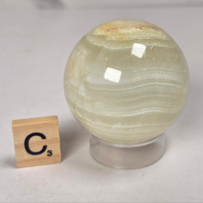 banded calcite spheres with green, brown and red banding on an acrylic stand with a scrabble tile against a marbled background
