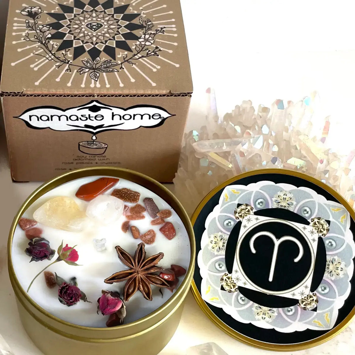 A decorative candle set from Namaste Home, featuring a gold tin candle filled with white wax, crystals, and spices, alongside its patterned box and a round lid with a black and white Aries zodiac symbol.