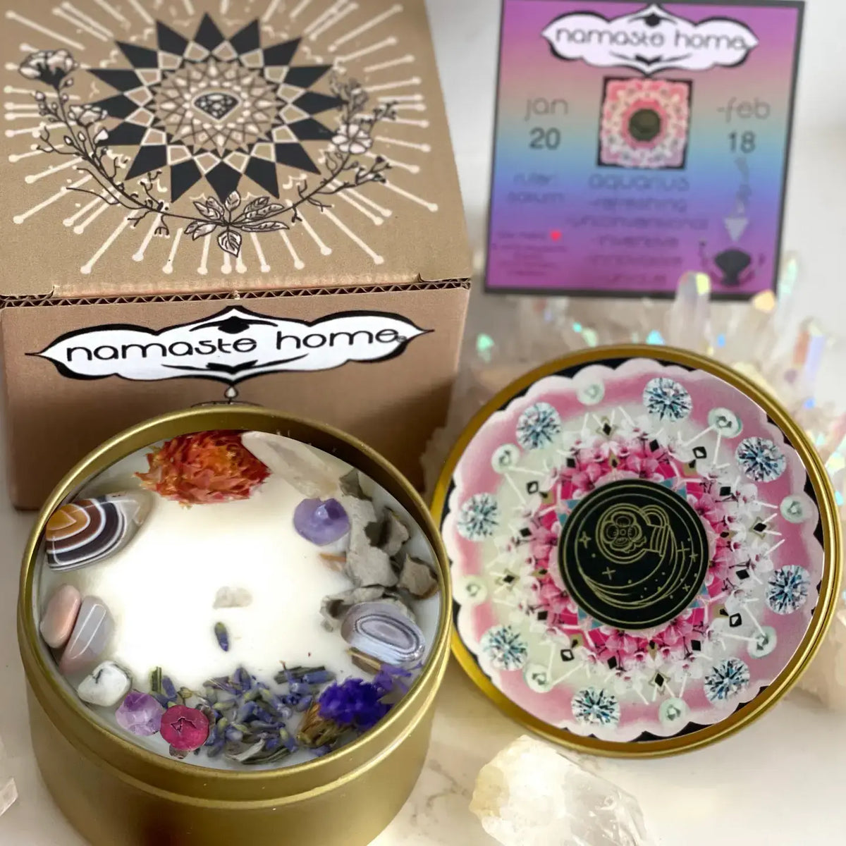 A decorative round candle tin shown open with white wax and embedded stones and dried flowers, alongside the lid placed beside it. A branded box labeled 'namaste home' and a small card are visible in the background on a light surface.