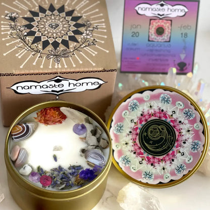 A decorative round candle tin shown open with white wax and embedded stones and dried flowers, alongside the lid placed beside it. A branded box labeled 'namaste home' and a small card are visible in the background on a light surface.