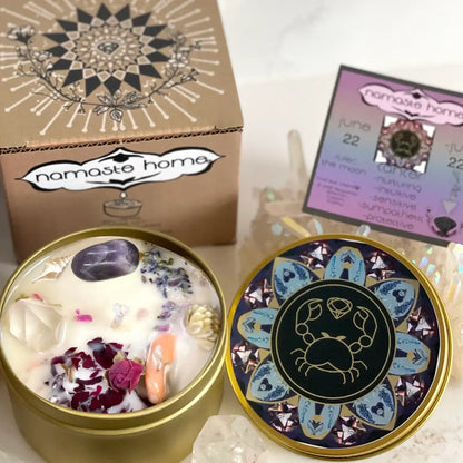 Namaste Home wax candle in a round gold tin, shown open with white wax, crystals, and dried flowers, alongside the patterned lid and branded box on a light marble surface.