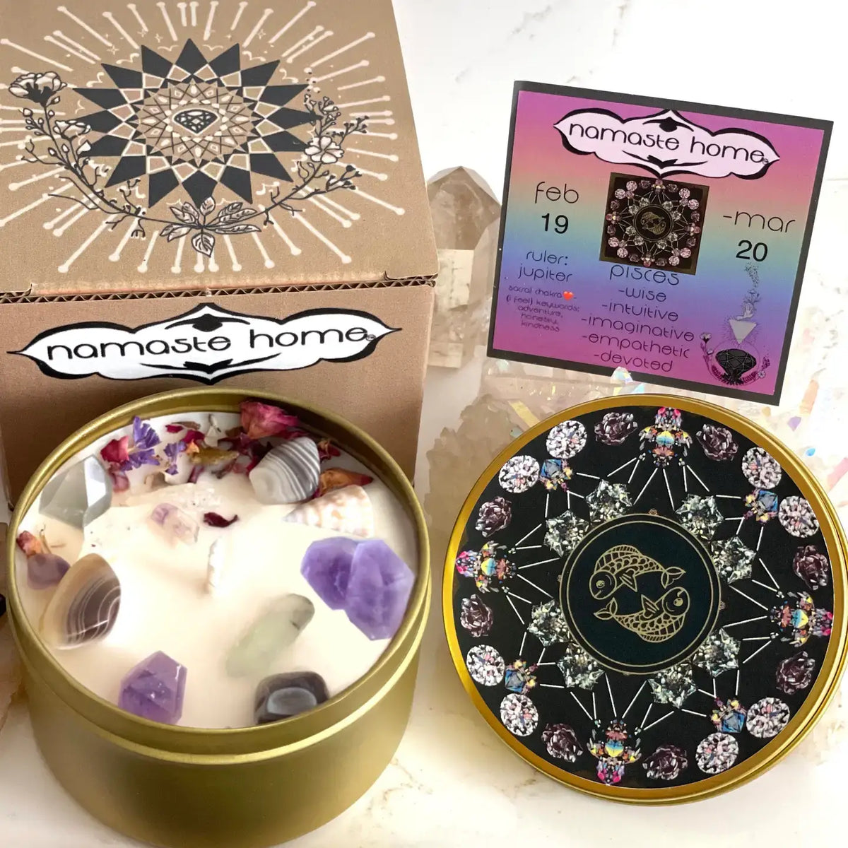 A brass round candle tin filled with white wax, purple crystals, and dried flower petals, shown alongside its kraft paper box and a colorful 'Namaste Home' calendar card on a light marble surface.