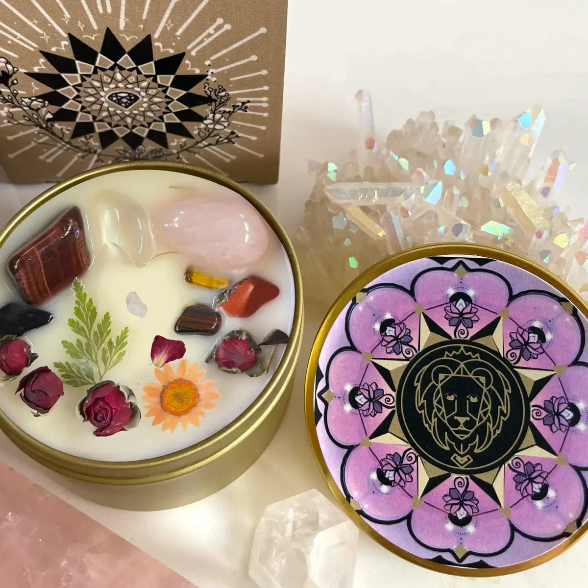 A round metal candle tin shown open, with white wax and embedded stones, dried roses, and a small flower. The lid beside it has a purple and black mandala-style design with a lion head in the center.