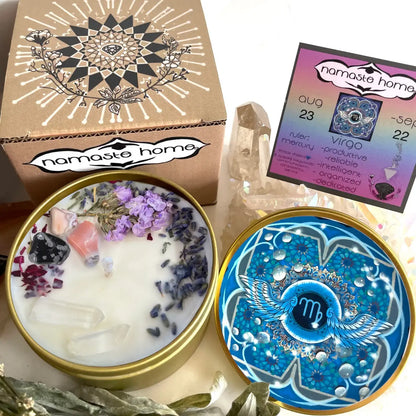 A decorative wax candle set from Namaste Home, featuring a round gold tin with white wax topped with crystals and dried lavender, its illustrated kraft box, the matching blue-and-white lid, a crystal cluster, and a printed card with astrological details.