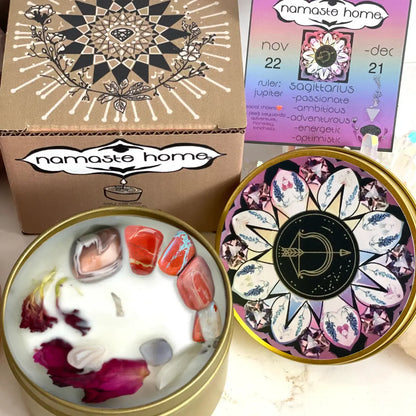 A decorative candle set from Namaste Home shown in its packaging and open tin. The set includes a kraft paper box with a black and white mandala design, a pastel-colored calendar card, and an open round tin filled with white wax, colorful crystals, and dried flower petals.