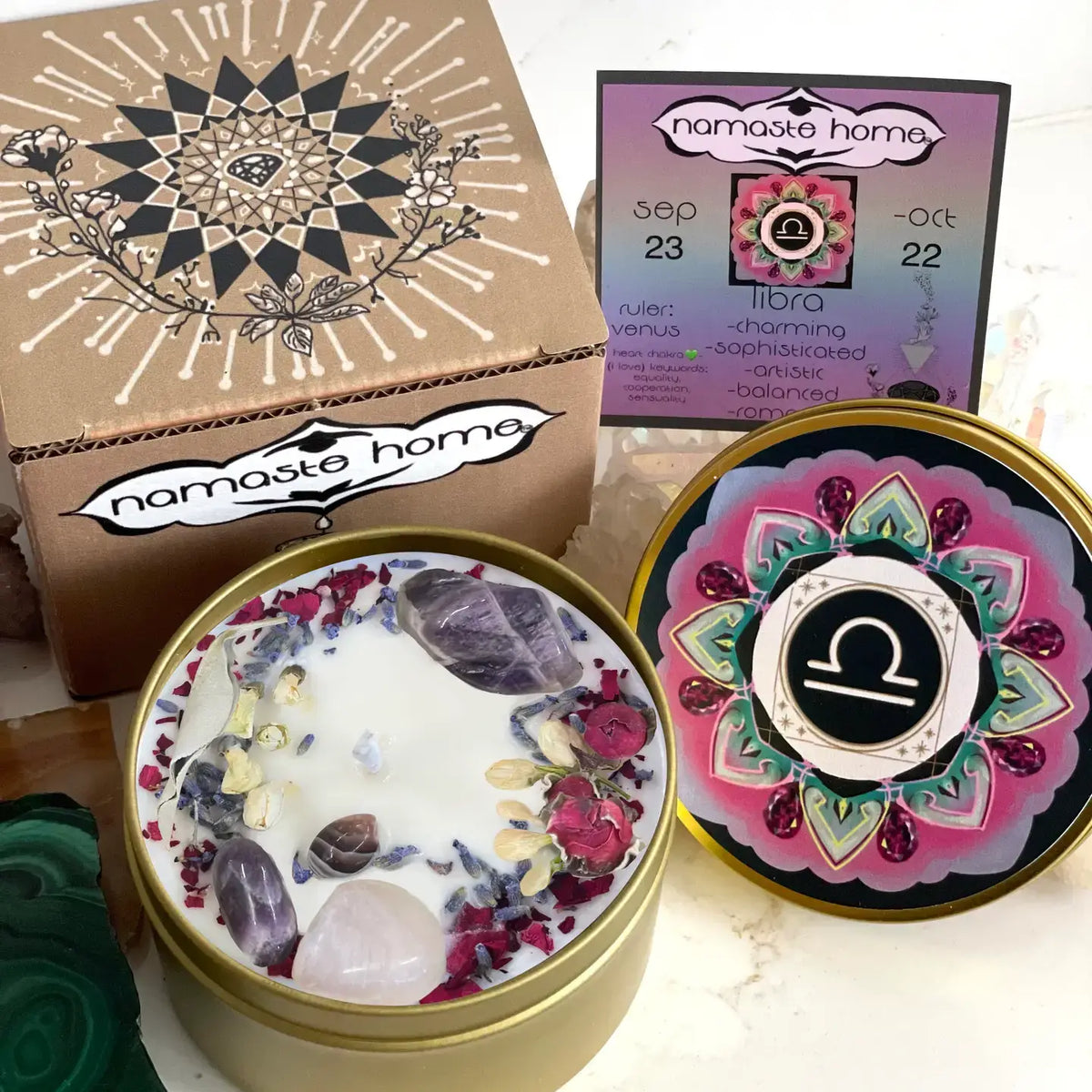 A spiritual candle set by Namaste Home, featuring a gold tin candle with white wax and embedded crystals, its lid shown next with a pink and gold mandala design, a kraft paper box with a black mandala, and a colorful calendar card.