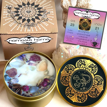 A decorative metal candle tin shown open with a white wax surface topped with crystals, shells, and dried flowers, alongside the printed lid design and branded box. A card reading 'namaste home' is also visible in the background.