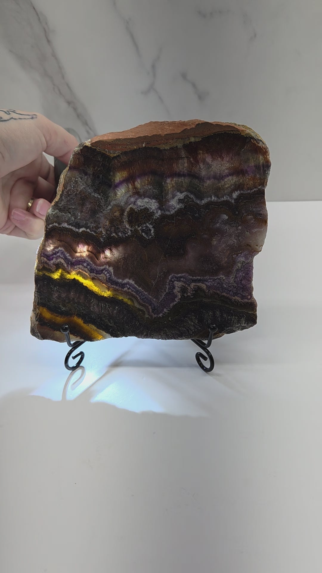 Banded crystal slice with multiple colors and inclusions on a stand against a marble background, being backlit