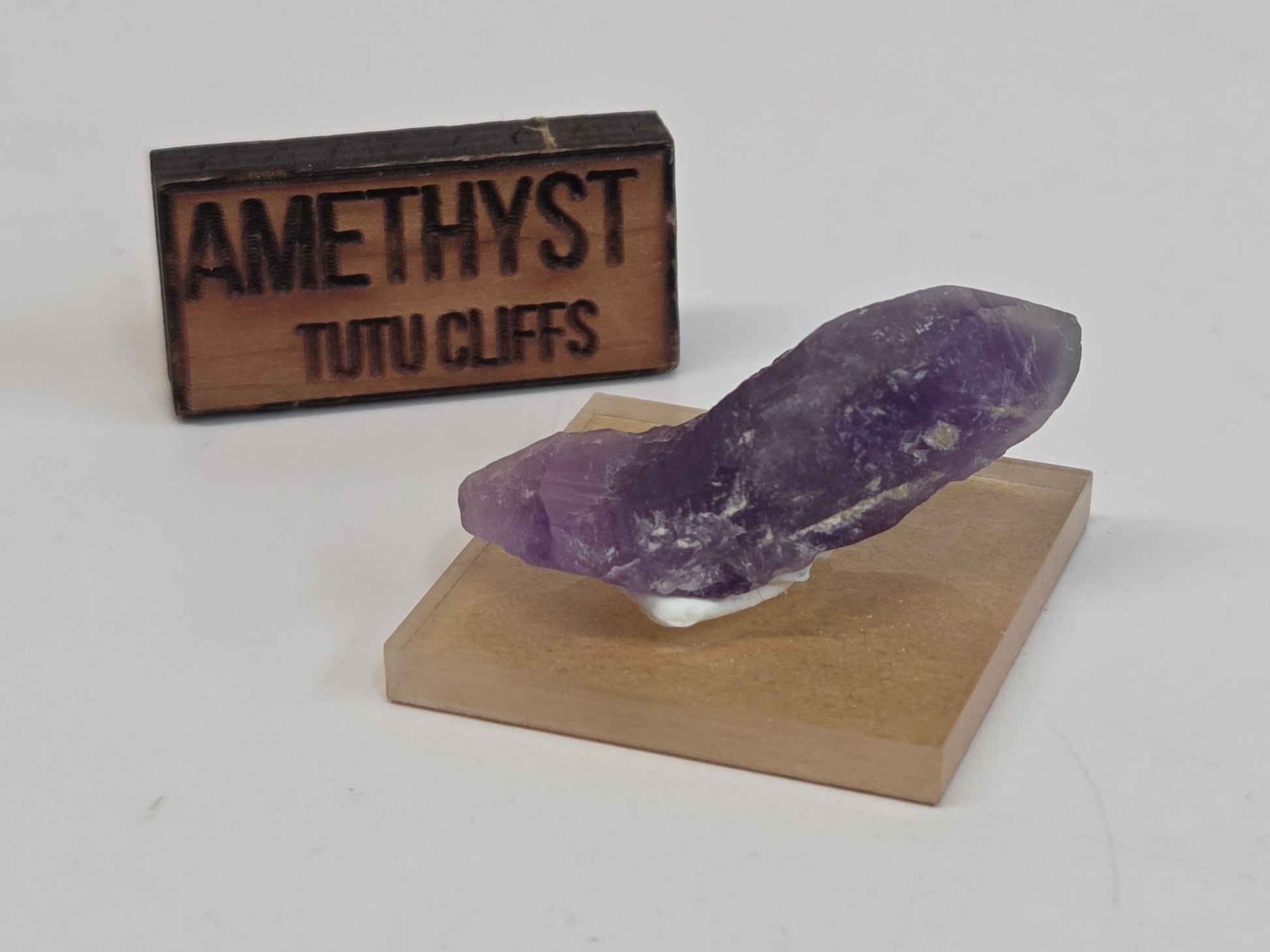 Amethyst crystal on a wooden stand with 'Amethyst Tutu Cliffs' sign on a white background